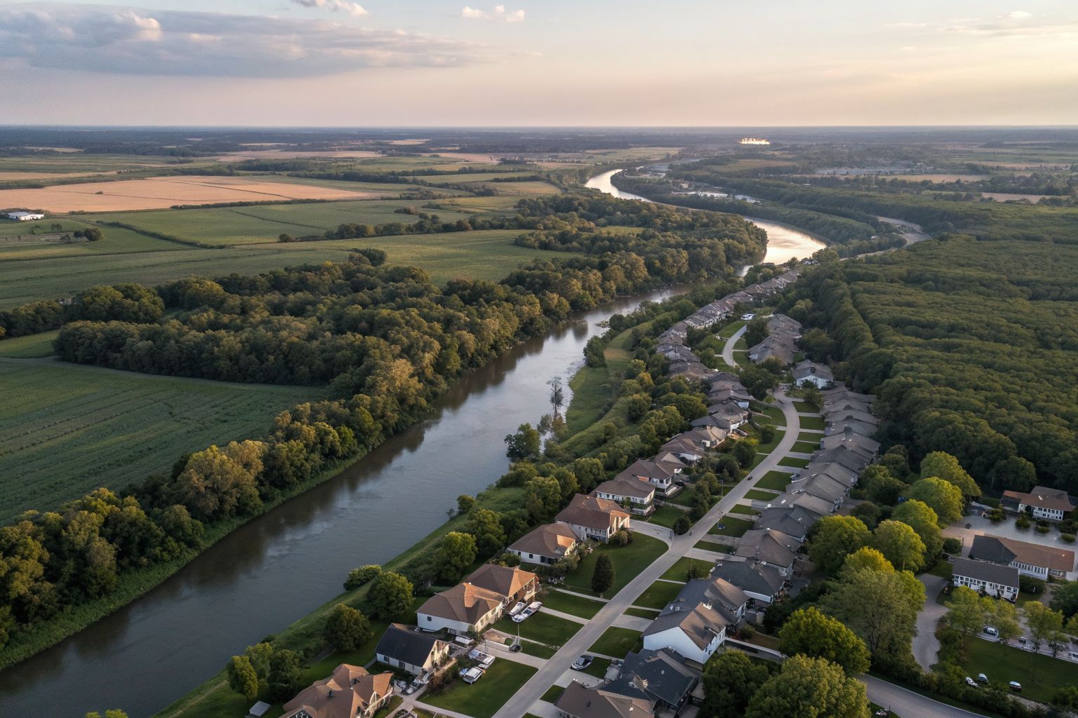 Aerial view of a suburb with shingle roofs beside a winding river and green farm fields